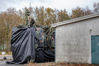 Militairen camoufleren een vrachtwagencabine met zwart zeildoek. De truck staat naast een grijs gebouw.