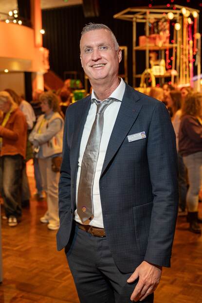 Man in pak kijkt lachend in de camera en staat in zaal vol mensen.