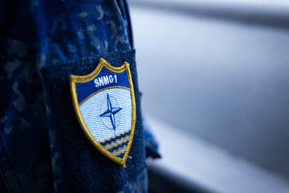 Links close-up van een mouwembleem van SNMG1. Rechts kijkt een marinevrouw door een verrekijker.