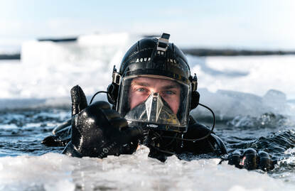 Een duiker met duikbril op steekt zijn hoofd bovenwater in een wak in het ijs.