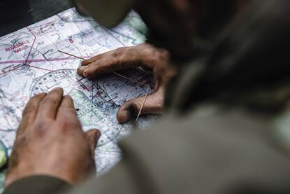 Links laat een marinier zich abseilend van een berg zakken. Rechts handen die een kaarthoekmeter gebruiken op een landkaart.
