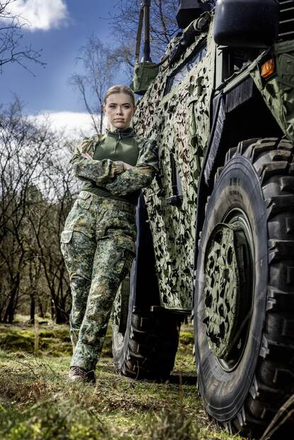 Een vrouwelijke militair leunt tegen een militair voertuig en kijkt in de camera.