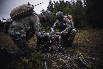 Twee militairen buigen zich over een gevonden kist en een parachute, in een open plek in het bos.
