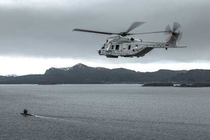 Een NH90 vliegt boven een fjord bij Bergen in Noorwegen met de onderzeeboot Dolfijn op de voorgrond.