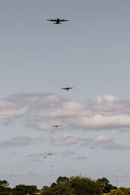 Een formatie met daarin dertien zichtbare toestellen vliegt achter elkaar door de bewolkte lucht.