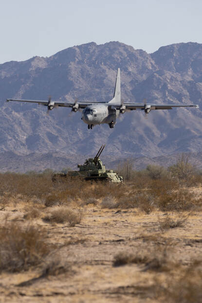 Een C-130 vliegt laag over het terrein waarin een pantserwagen tegen luchtdoelen staat opgesteld.