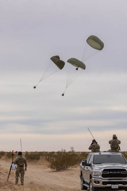 Militairen op de grond en in een voertuig kijken naar de parachutes met bevoorrading in de woestijn.