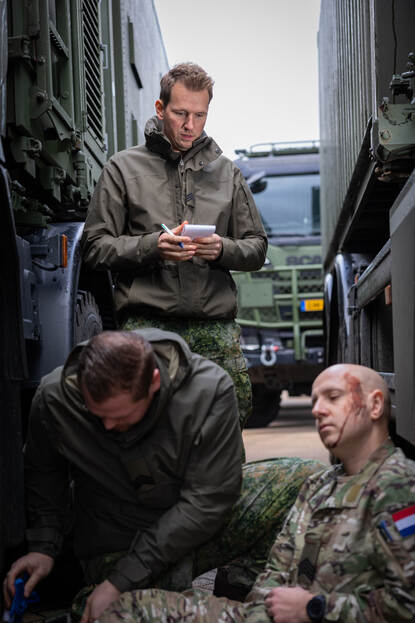 Tussen twee militaire vrachtwagens zit een militair met een hoofdwond. Een collega verleent eerste hulp. Sergeant Tom staat ernaast te kijken met een notitieboekje in de hand.