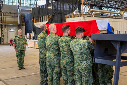 Een lijkkist wordt aan weerszijden vastgehouden door vier personen. Daarop ligt een Nederlandse vlag. De commandant van de ploeg staat enkele meters verderop en houdt het geheel in de gaten.