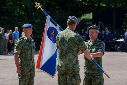 Een militair met een vlag in zijn handen staat tegenover een militair die de vlag in ontvangst gaat nemen. Een derde militair kijkt aan de zijkant toe.