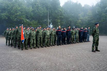 Militairen en brandweerlieden op een rij. Een ervan heeft een vlag in de hand.
