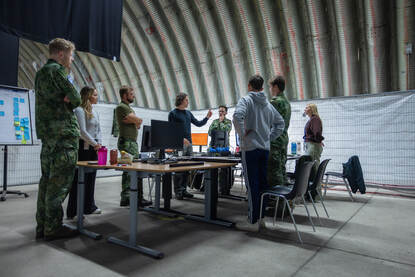 AMilitairen en burgers staan rondom een tafel met daarop computers. Een van de burgers voert het woord en maakt daarbij een wijzende beweging.