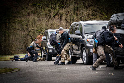 Drie zwarte auto’s staan naast elkaar opgesteld, daarachter zwaarbewapende BSB’ers die dekking zoeken. Vanachter de auto’s vandaan schieten ze op hun doel.