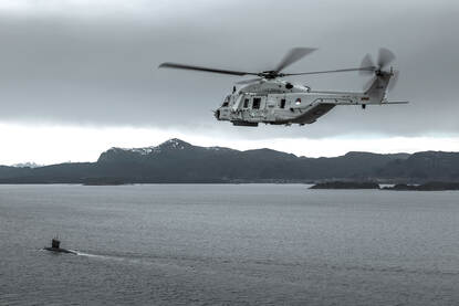 NH90-helikopter boven zee bij bergachtige kustlijn met een onderzeeboot.