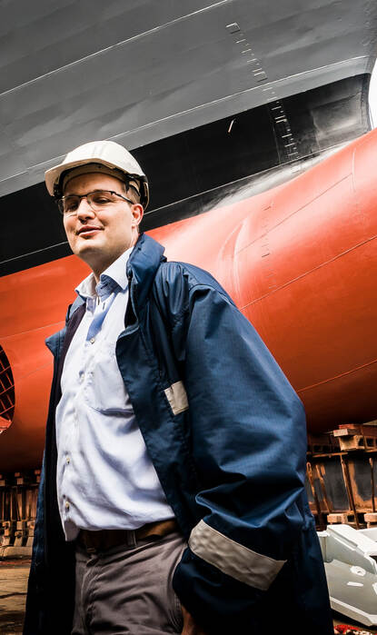 Roy met veiligheidshelm en werkjas staat in een scheepswerf voor de rood geschilderde boeg van een groot schip dat op steunblokken rust.